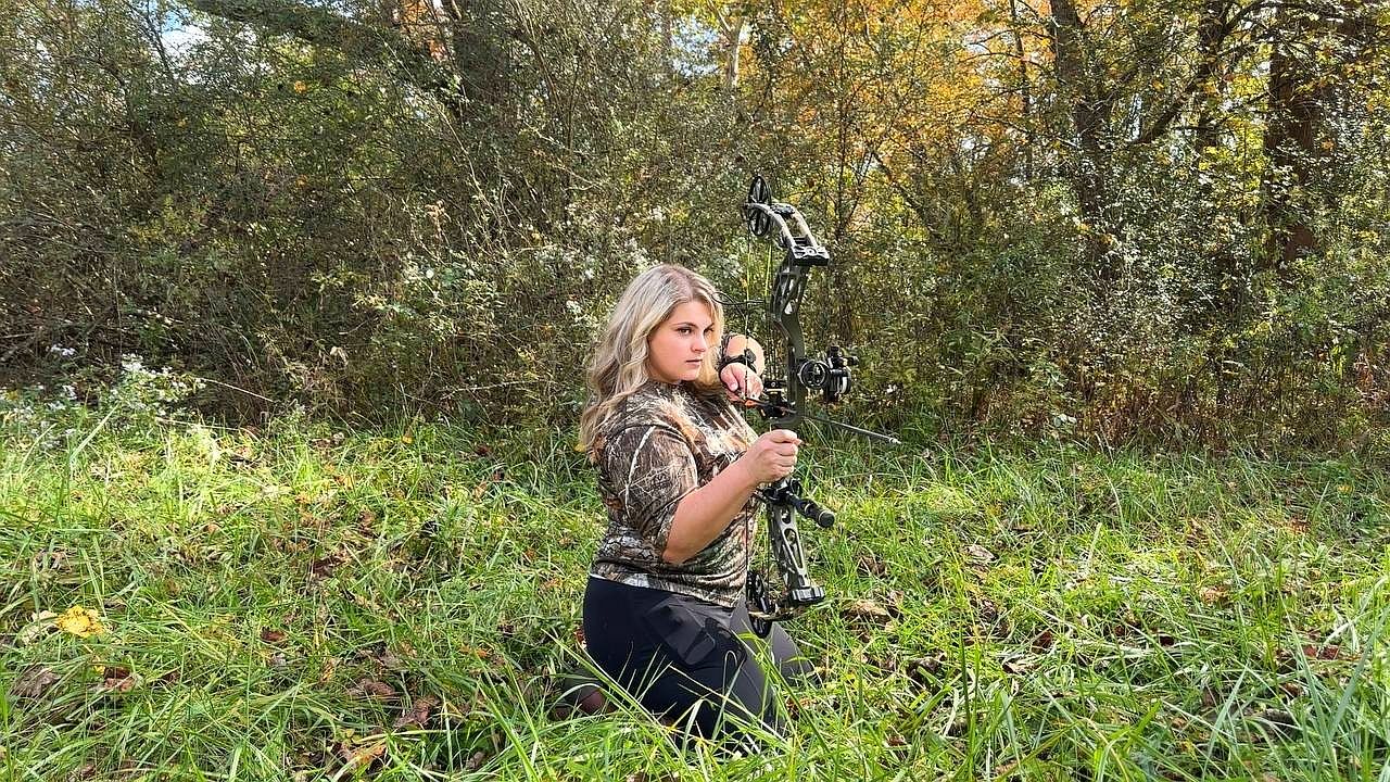 Bree kneeling in the grass with her compound bow, wearing a camo shirt